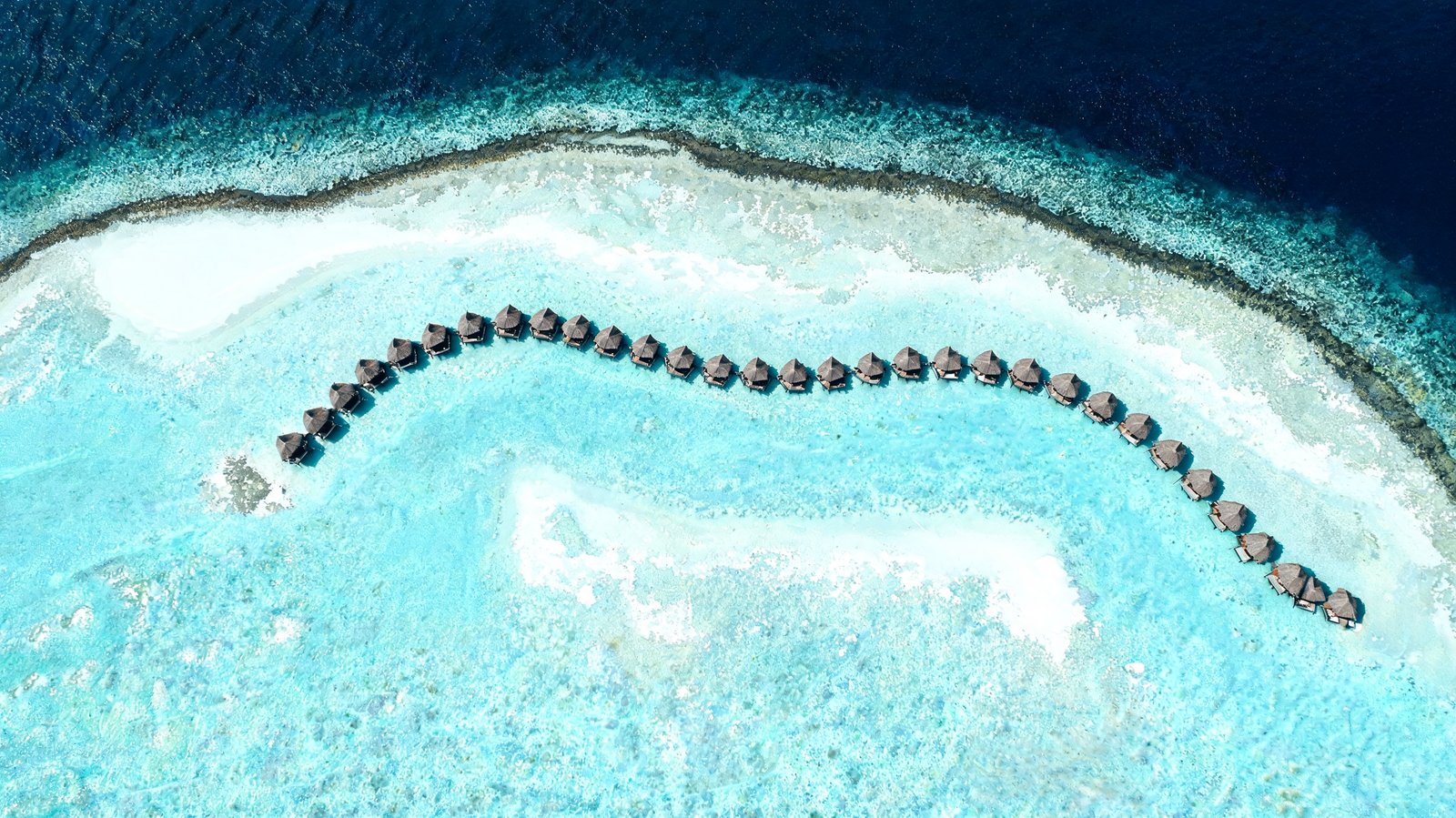 A curving line of overwater villas above turquoise lagoon shallows, seen from directly overhead, Maldives