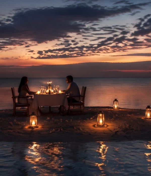 A couple at a candlelit table set on the sand at dusk, lanterns along the waterline