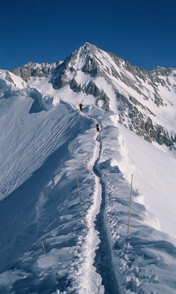 A boot-packed trail climbing a snow ridge toward Highland Bowl with two distant skiers carrying skis
