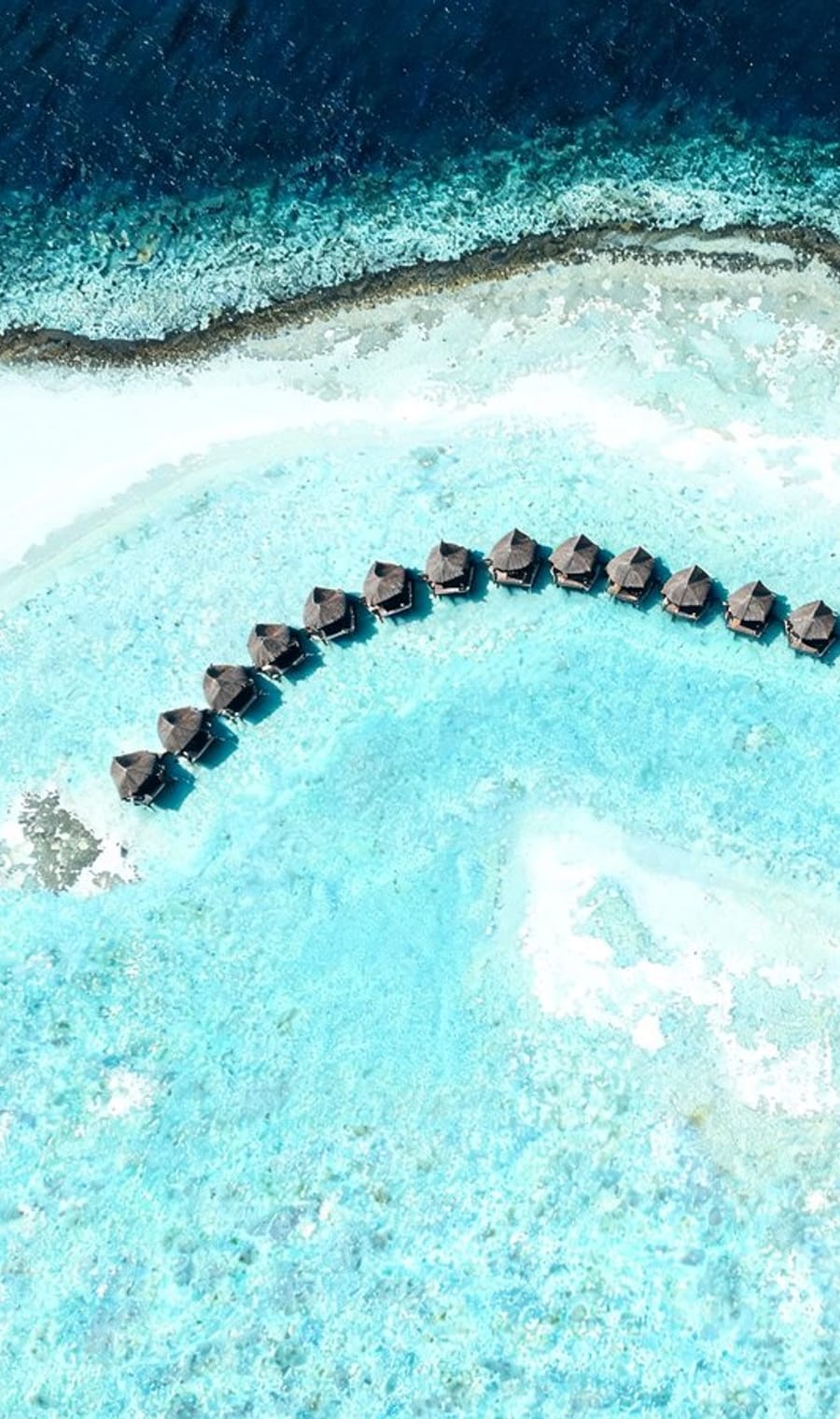An aerial view of overwater villas curving across the turquoise shallows of a Maldivian atoll