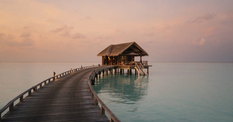 A single thatched overwater villa at the end of a curving jetty above still lagoon water at dusk